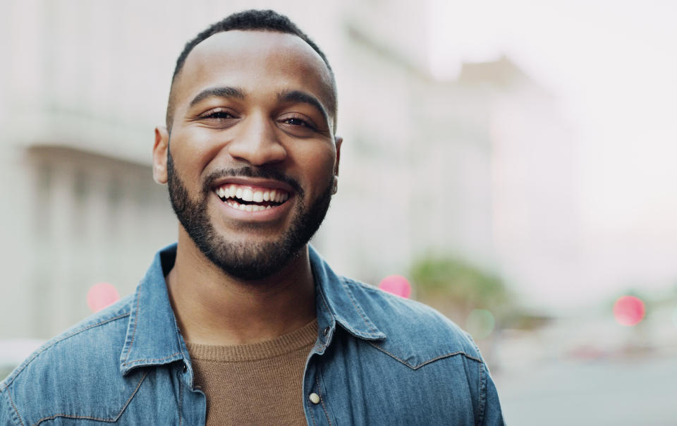 A man with a beard and mustache smiles at the camera while wearing a casual denim jacket over a shirt