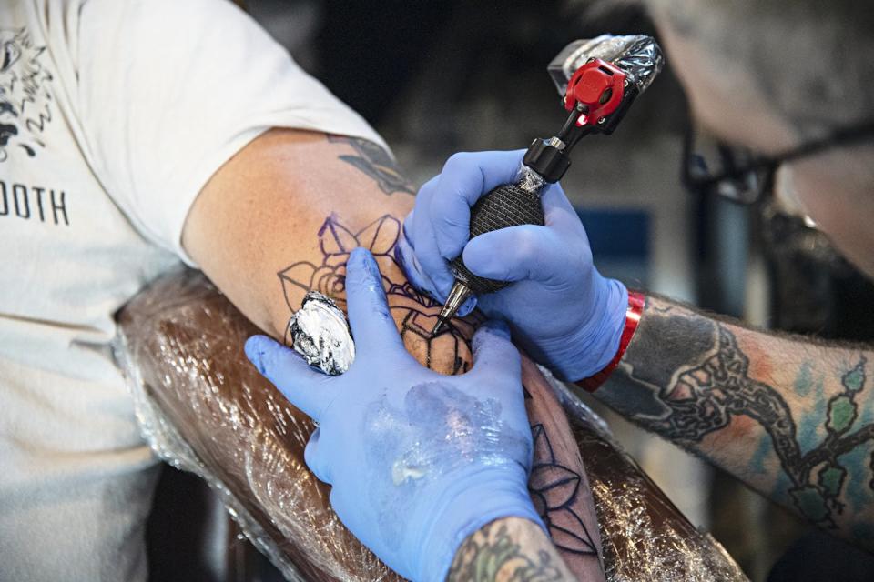 A tattoo artist wearing latex gloves holding a tattooing needle inks a geometric design on an arm.