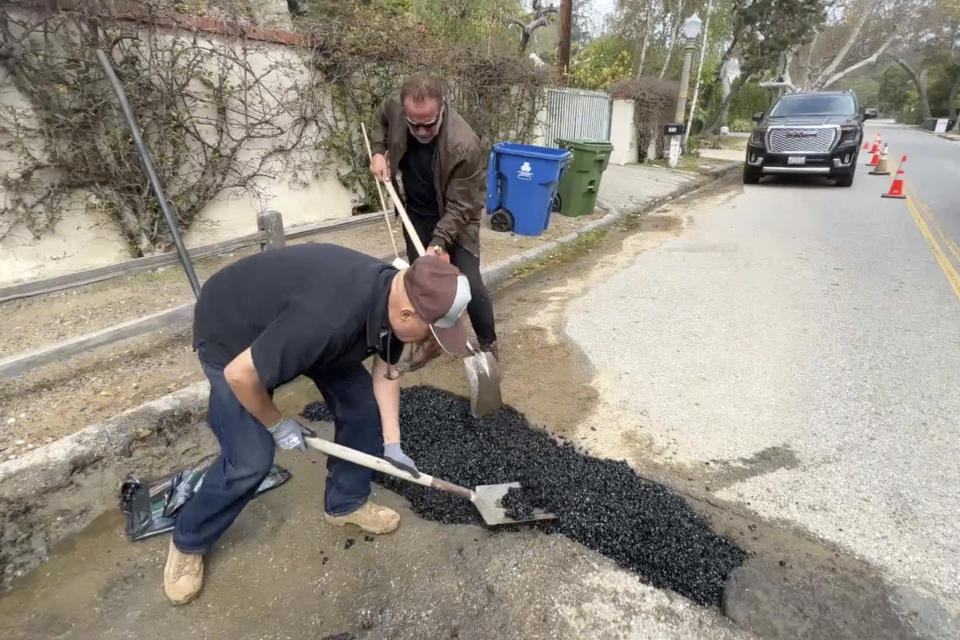 This video still image provided by The Office of Arnold Schwarzenegger, shows former California Governor Arnold Schwarzenegger, center back, repairing a pot hole on a street in his Los Angeles neighborhood on Tuesday, April 11, 2023. Fed up by an enormous pothole in his neighborhood, Schwarzenegger picked up a shovel and filled it himself. (The Office of Arnold Schwarzenegger via AP)