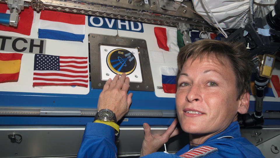 peggy whitson wearing a blue flight suit sticks a mission patch among different flags.