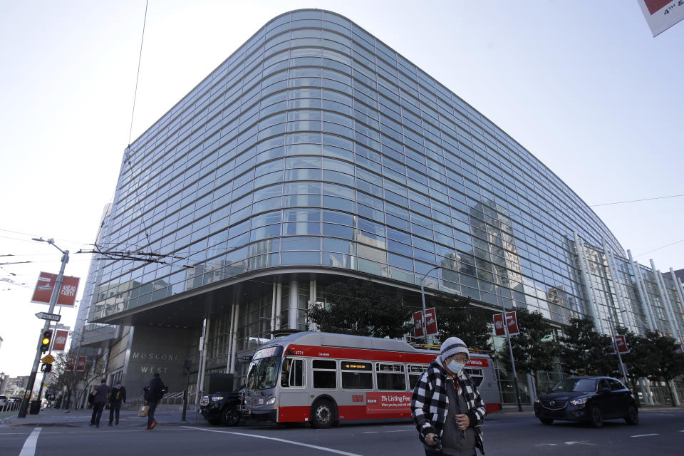 A woman wears a mask while crossing the street in front of Moscone Center West in San Francisco, Thursday, April 2, 2020. Officials are setting up 400 beds at the center to house homeless people who are currently in shelters to allow for more social distancing during the coronavirus pandemic. (AP Photo/Jeff Chiu)