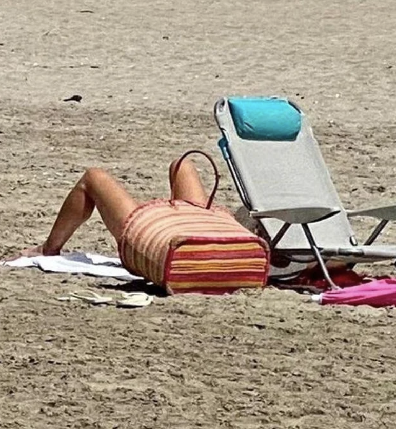 Person laying on a beach with their head obscured by a large striped bag, next to a folding chair with a teal cushion