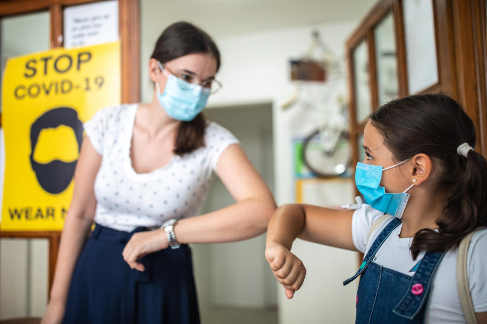 Teacher greeting school children with elbow bump before they enter school, they are wearing protective face masks for protection against virus during covid-19 pandemic, poster about virus prevention is on doors