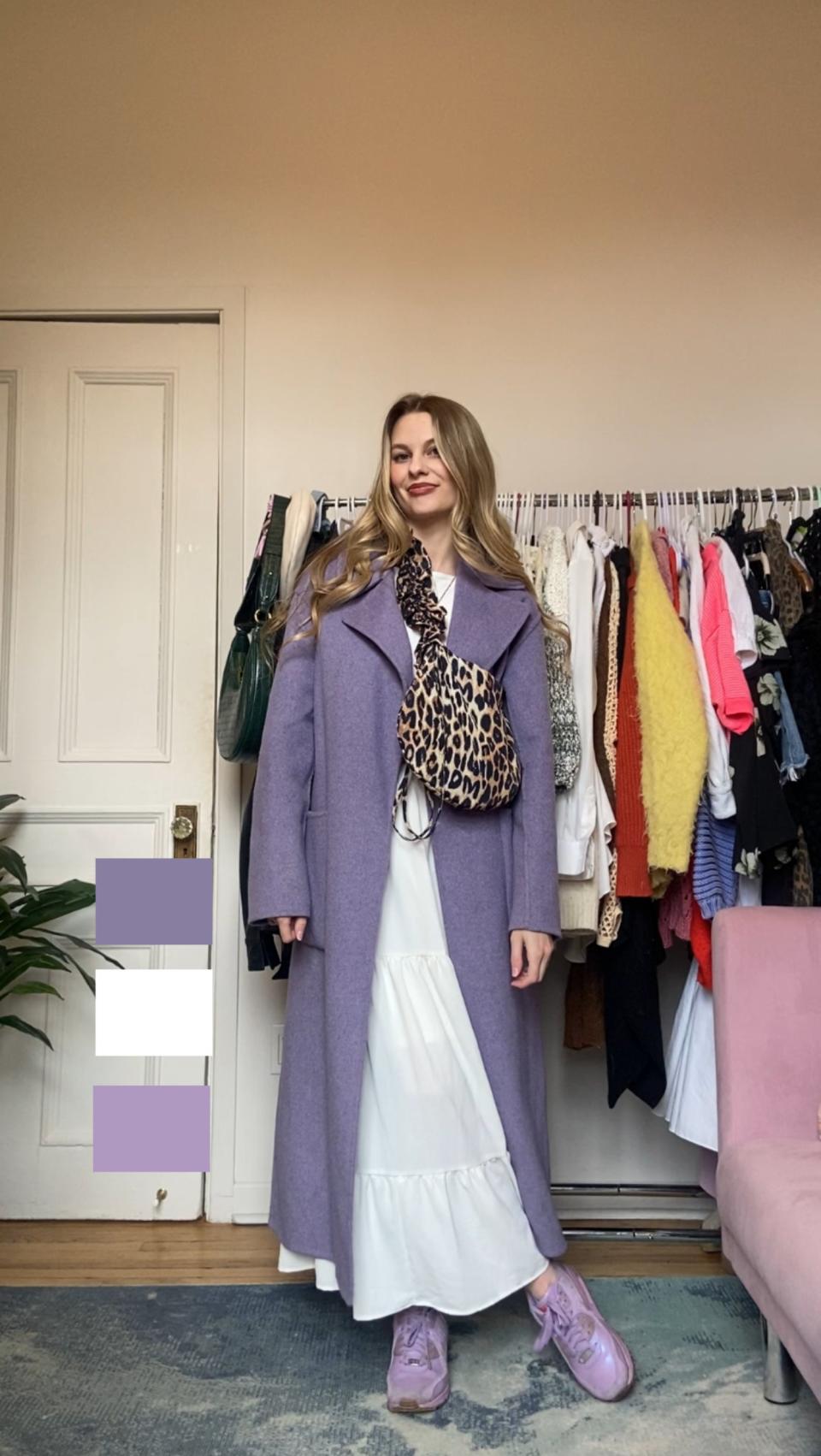 Woman in a long purple coat, leopard print top, and white skirt standing in a room with clothes rack behind