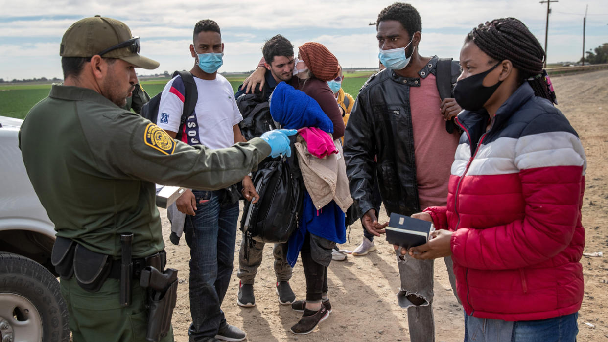 A uniformed Border Patrol agent talks with a group of people wearing facemasks on a dirt road.