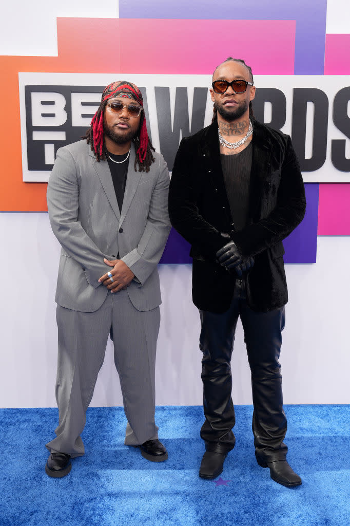 Leon Thomas III and Ty Dolla Sign in stylish attire pose at an awards event. Both wearing sunglasses, one is in a gray suit and the other in a black velvet blazer with leather pants