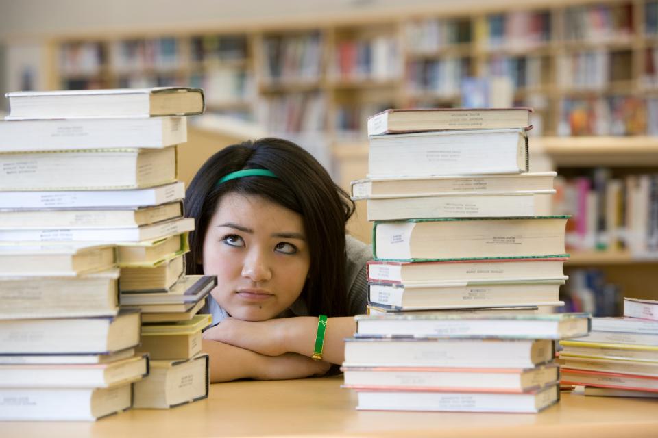 A woman sits at a table in a library, looking overwhelmed while surrounded by tall stacks of books