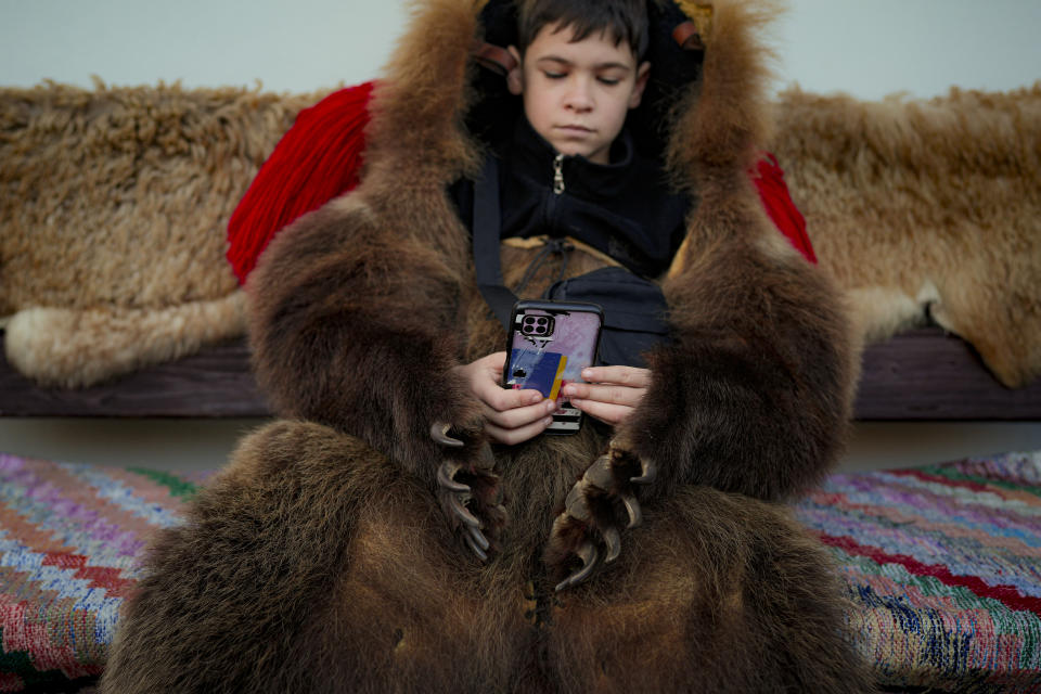 A boy looks at a mobile phone while waiting, dressed in a bear fur costume, to take part in a parade showcasing regional winter traditions in Comanesti, northeastern Romania, Friday, Dec. 30, 2022. The custom originated in pre-Christian times, when dancers wearing colored costumes or animal furs went village households, singing and dancing to ward off evil. (AP Photo/Vadim Ghirda)