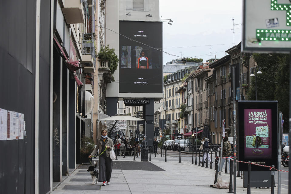 People walk past a giant screen streaming Versace fashion live show, during the Milan's fashion week in Milan, Italy, Friday, Sept. 25, 2020. (AP Photo/Luca Bruno)