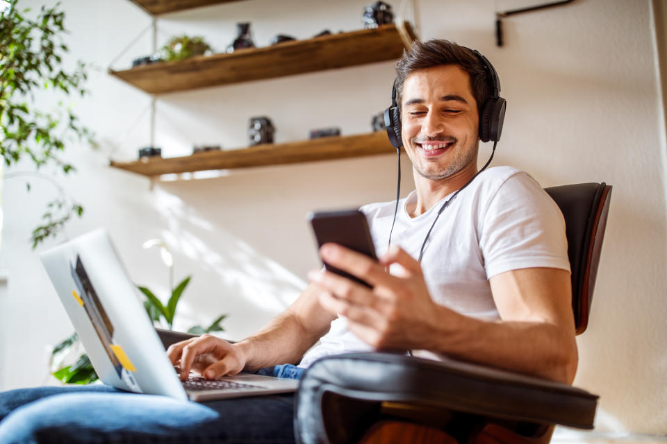Young man wearing headphones listening to music from mobile phone while working on laptop at home.