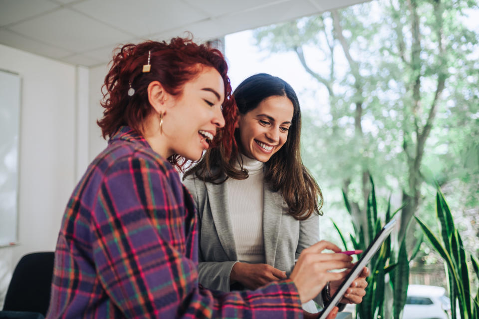 A young trendy woman is holding a tablet and pointing at it while giving investing tips for investing in fractional shares to a colleague, family member or friend