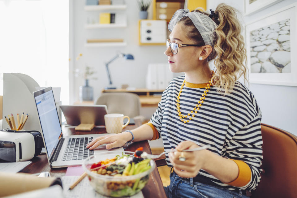 A woman wearing a striped top and orange bead necklace eats a salad bowl while she works on her laptop at home