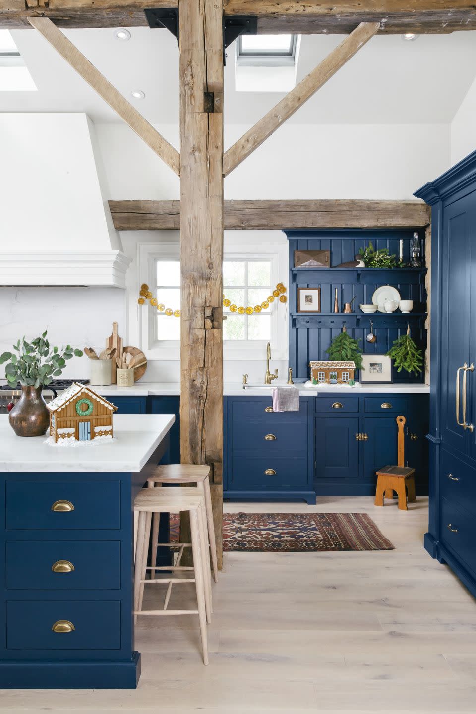 large dark blue kitchen island with drawers, gold toned drawer pulls, white counters, and blond wood stools for seating