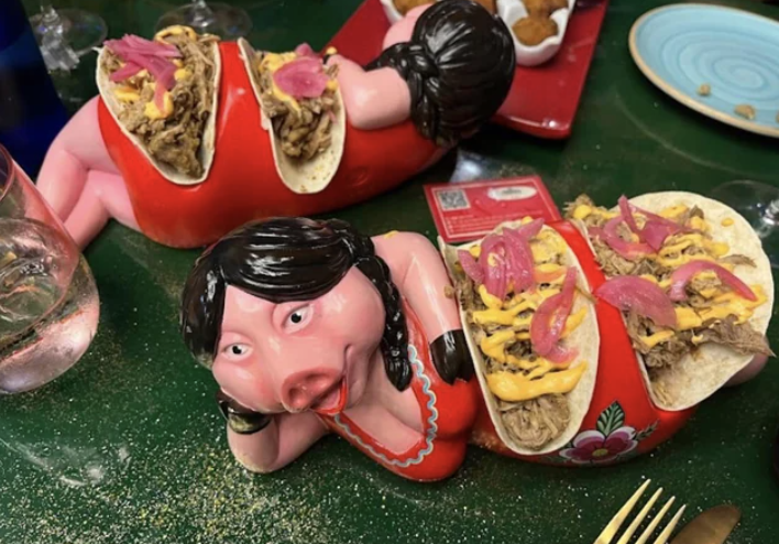 Three ceramic pig figures hold tacos on their backs in a creative serving dish, set on a green table with other food items visible