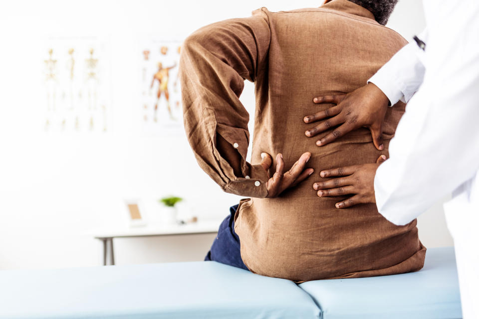 A patient with back pain, wearing a brown shirt, is being examined by a healthcare professional in a white coat, who is placing hands on the patient's back