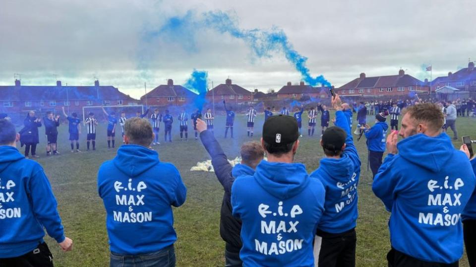 People on a football pitch holding blue smoke canons and wearing blue hoodies that say 'RIP Max and Mason'