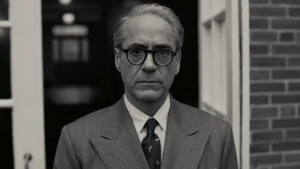 Robert Downey Jr. in glasses and a suit standing near a doorway in a black and white scene from Oppenheimer
