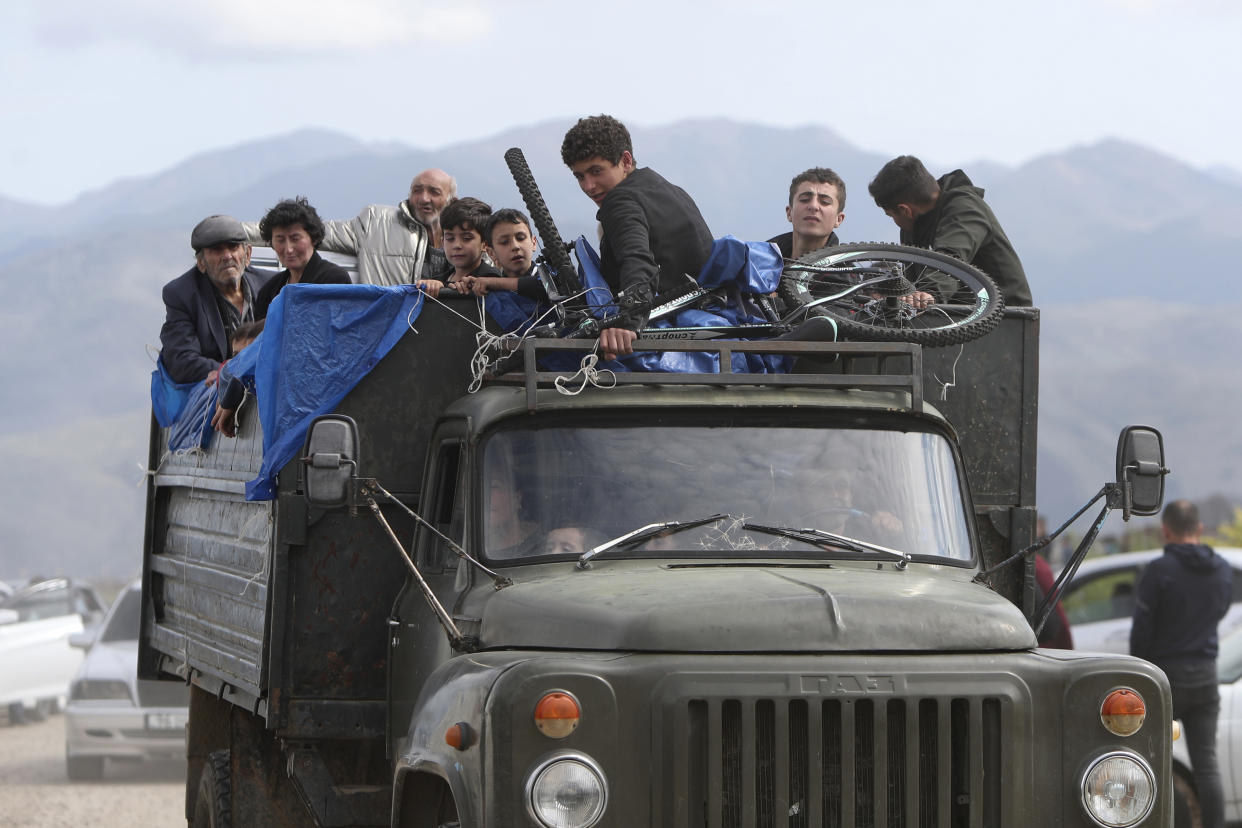 A military style truck with a cracked windshield and an open bed holds over a dozen people along with a bicycle tied to the roof of the cab.