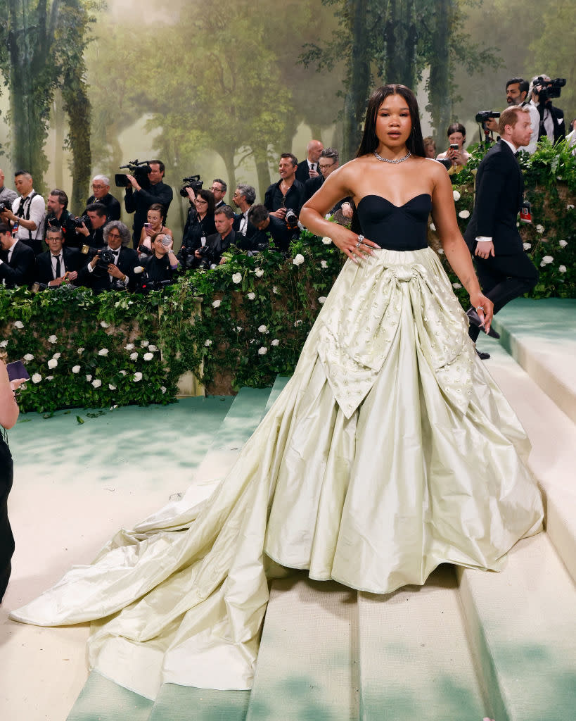 Person in a black sweetheart neckline top and large cream skirt with a train, standing on steps with photographers in the background