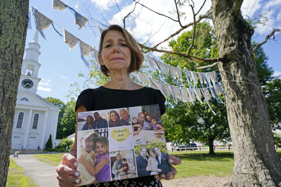 Widow Marcy Jacobs holds family photos as she stands under flags with names of people, including her husband, Keith Jacobs, who have died from COVID-19, outside the First Congressional Church, Thursday, June 17, 2021, in Holliston, Mass. The flags are part of the COVID Art and Remembrance project spearheaded by Marcy's daughter, Jaclyn Winer. (AP Photo/Elise Amendola)