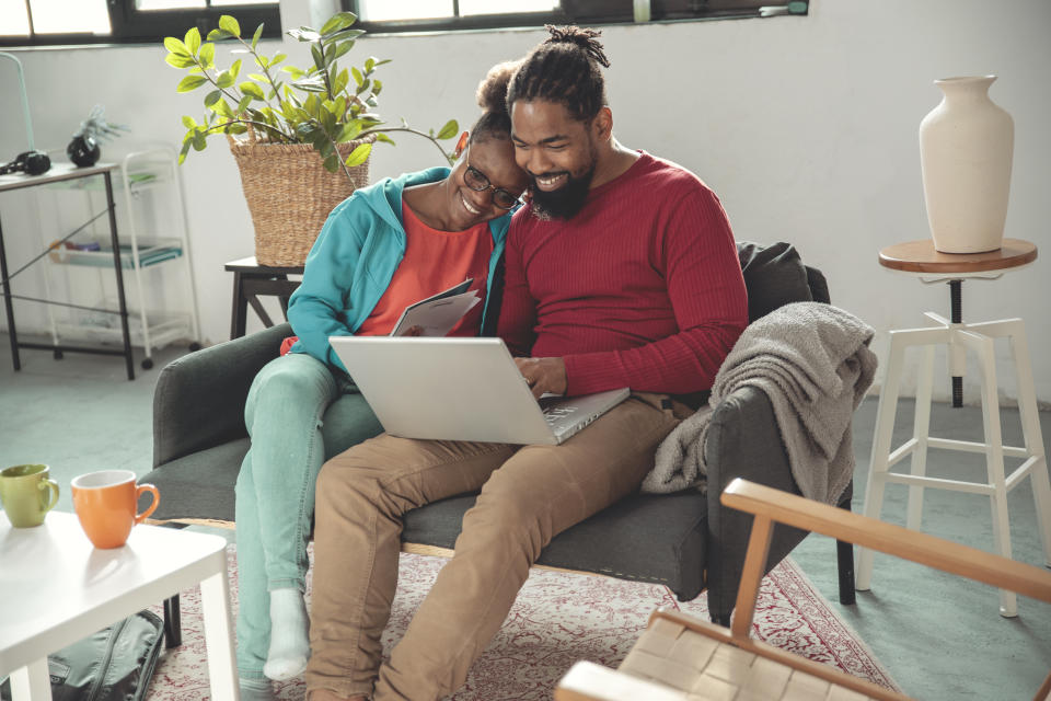 A couple sitting on a sofa, smiling and looking at a laptop together. A plant and various coffee mugs are on a table nearby