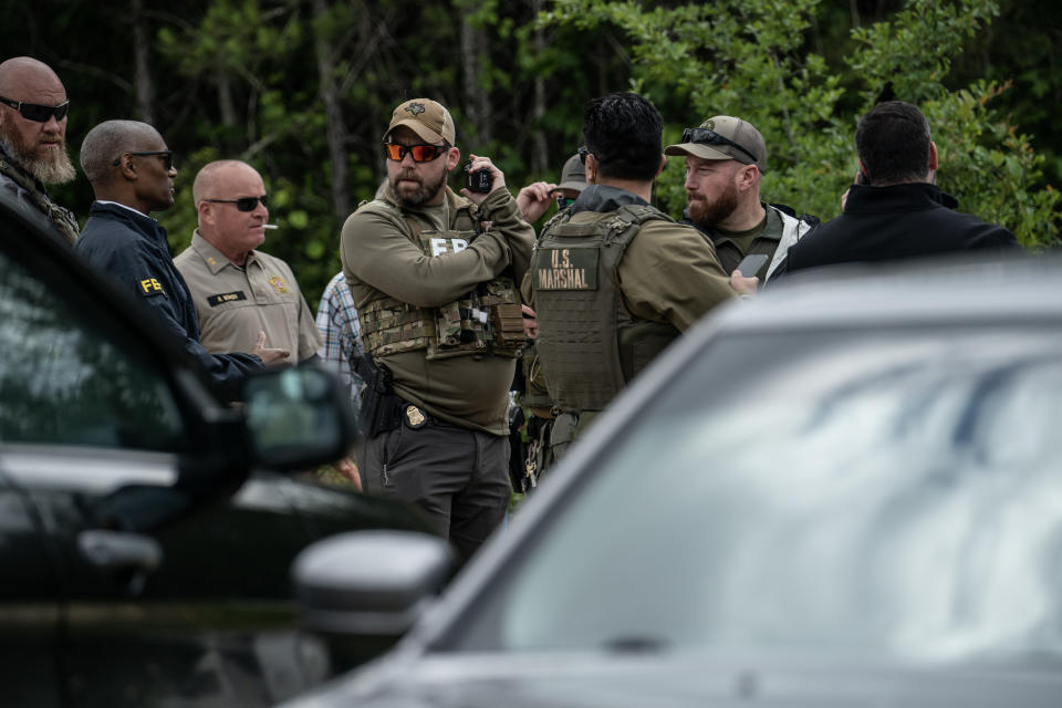 A half dozen law enforcement members, some with FBI or U.S. Marshal emblazoned on their clothes, stand near several cars and in front of a row of trees.