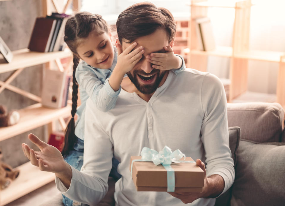 Cute little girl is giving her handsome father a gift box. Both are sitting on couch at home and smiling