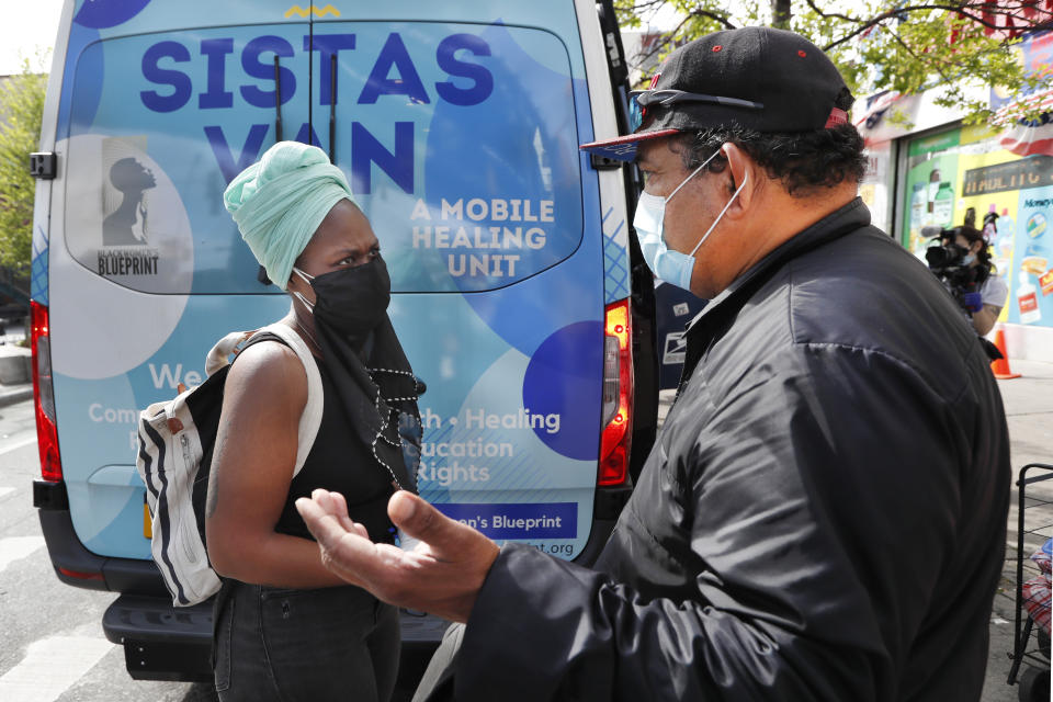 Sistas Van volunteer Sequaña Williams-Hechavarria, left, listens as Francisco Martinez, who lost his job due to an injury and now sells face masks from a cart on the street, makes his case for help after passing by a table the group had set up at Brooklyn's Broadway Triangle, a busy intersection and transit hub in the middle of Brooklyn, amid the coronavirus outbreak, Tuesday, May 19, 2020, in New York. Twice weekly, Sistas Van workers and volunteers deliver resources to some of the hardest hit areas of New York city, often immigrant communities and communities of color. (AP Photo/Kathy Willens)