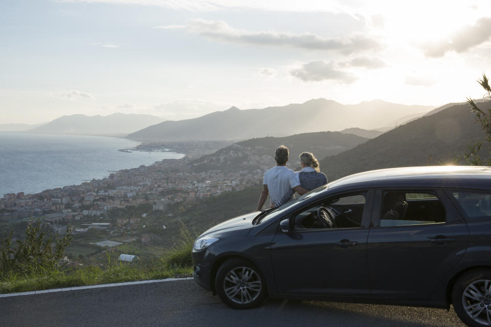 Couple relax against car, look past village to sea