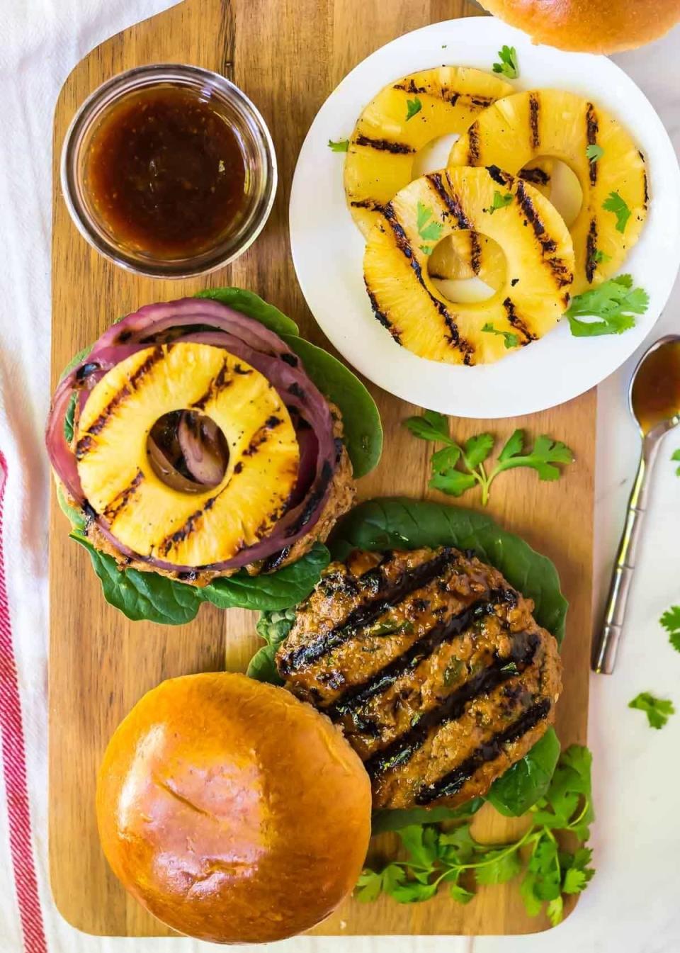 A grilled burger open-face style with spinach, grilled onions, and a grilled pineapple ring on a wooden board. The plate next to it has extra grilled pineapple slices