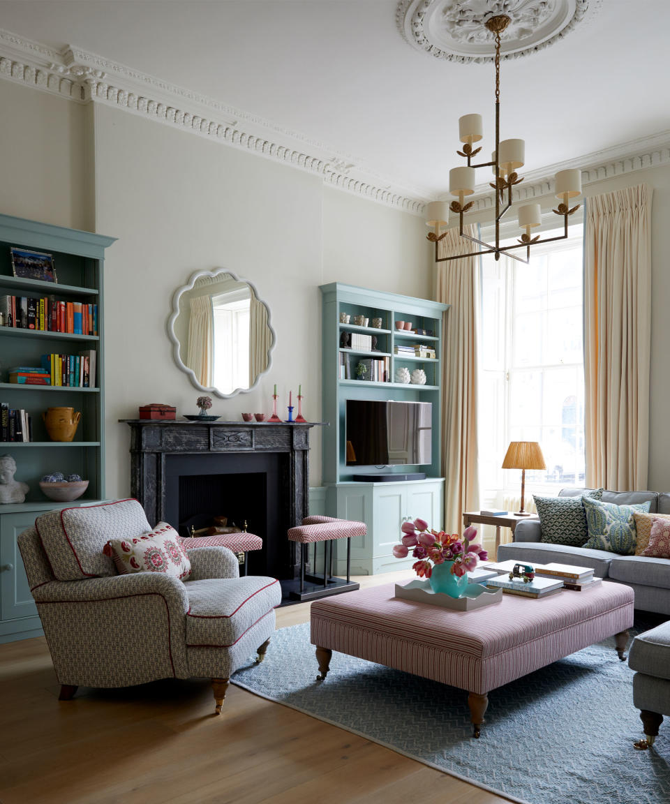 Living space with painted blue built in shelving either side of a black fireplace, rounded mirror with white scalloped edge, patterned armchair with red trim facing a large, red striped ottoman, large blue rug over wooden floorboards, metallic hanging pendant with seven cream shades, cream curtains