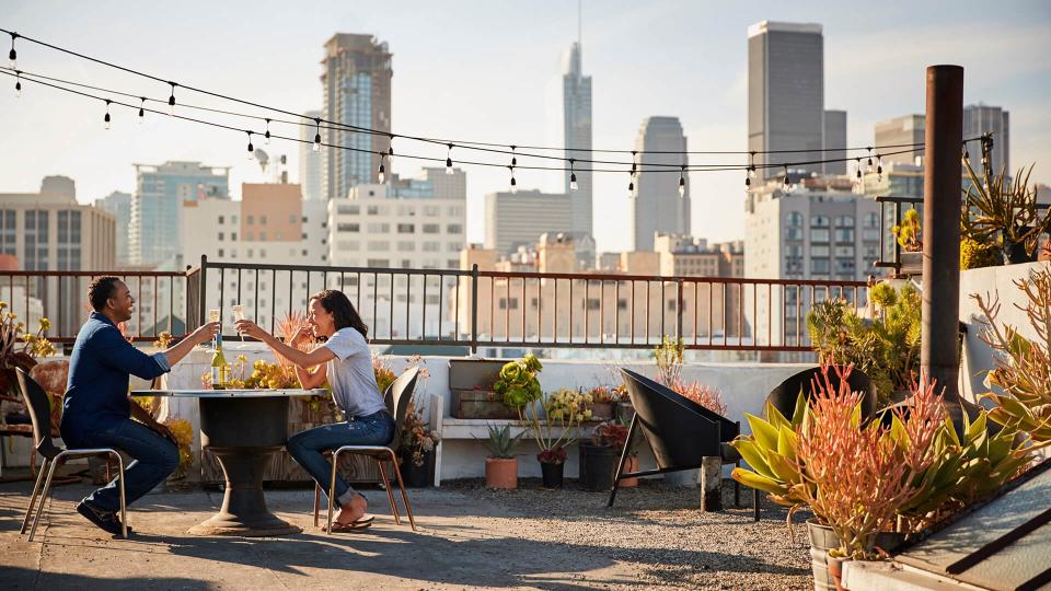 Couple Drinking Wine And Making Toast On Rooftop Terrace With City Skyline In Background.