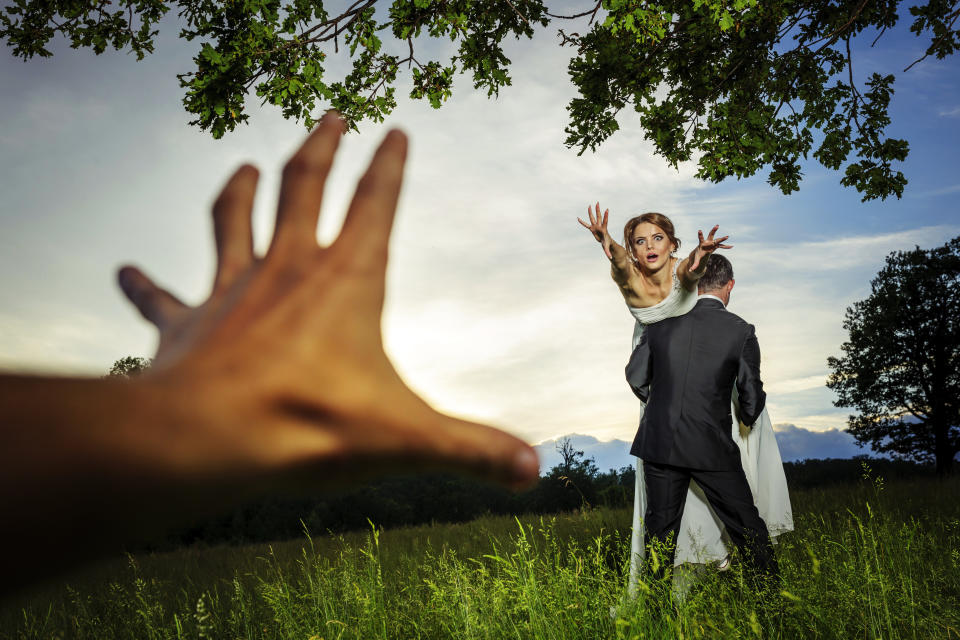 Newlywed couple in a field, bride reaching toward a hand in the foreground while being carried by the groom