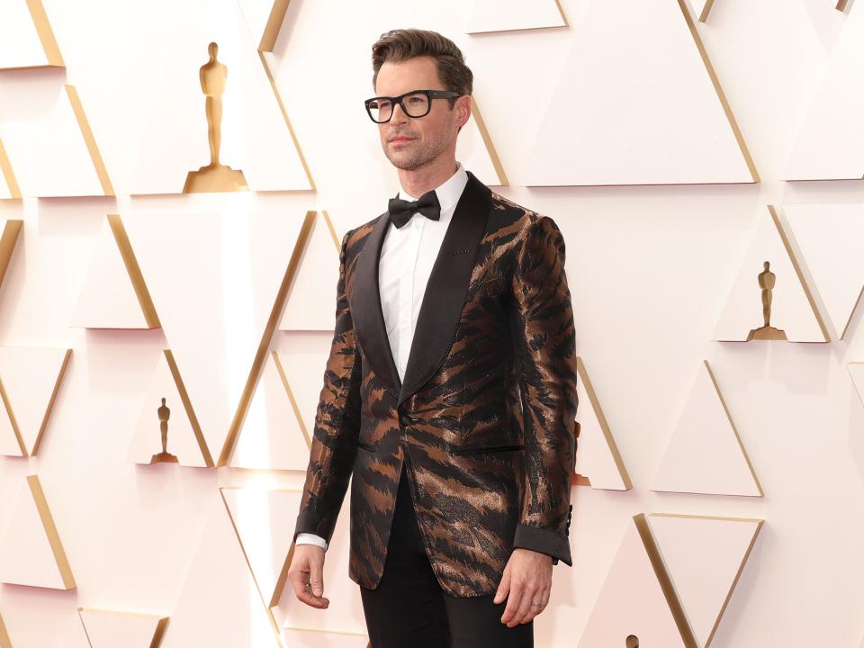 Brad in a shiny brown zebra print blazer with brown-black lapels, pants, and a bow tie. He's wearing a white button down and glasses.