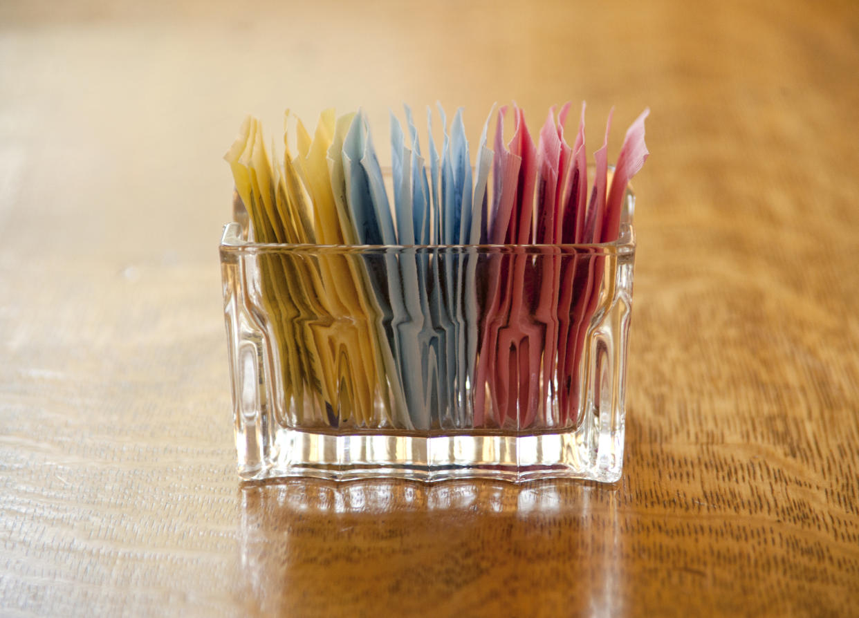 A glass container with packets of sugar substitutes on a table.