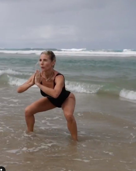 A photo of Elsa Pataky wearing a black swimsuit doing squats in the shallow water at the beach.