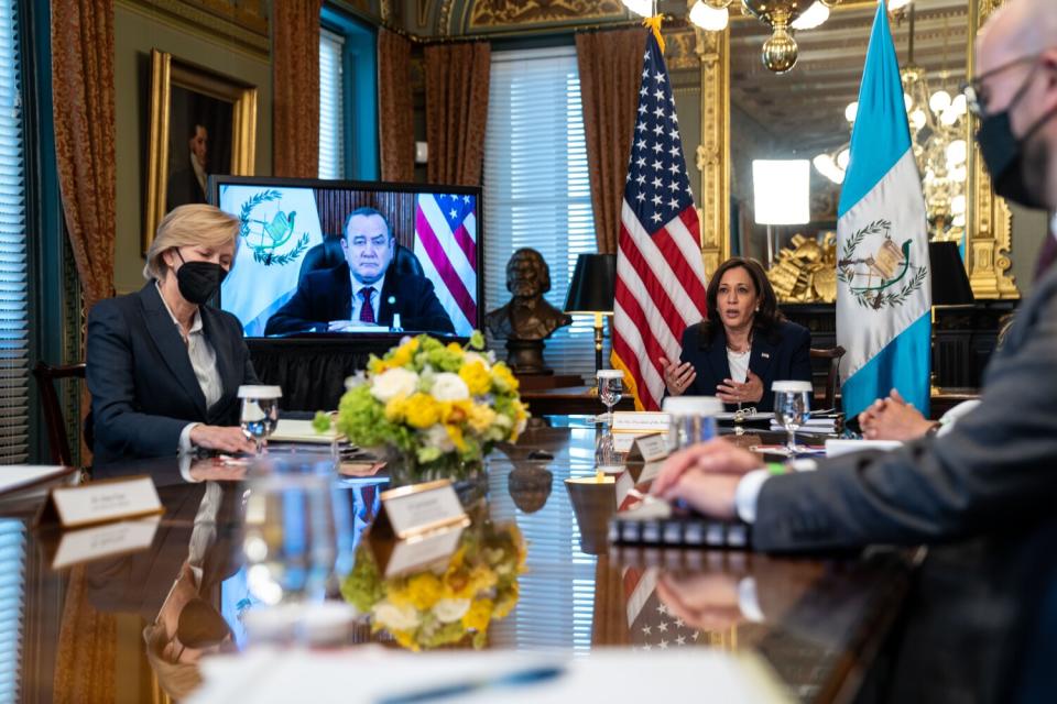 Vice President Kamala Harris sits at a table with others while Alejandro Giammattei appears on a TV screen nearby.