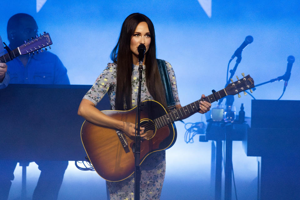 Kacey Musgraves performs on stage, playing an acoustic guitar and wearing a floral dress. Musicians and instruments are visible in the background