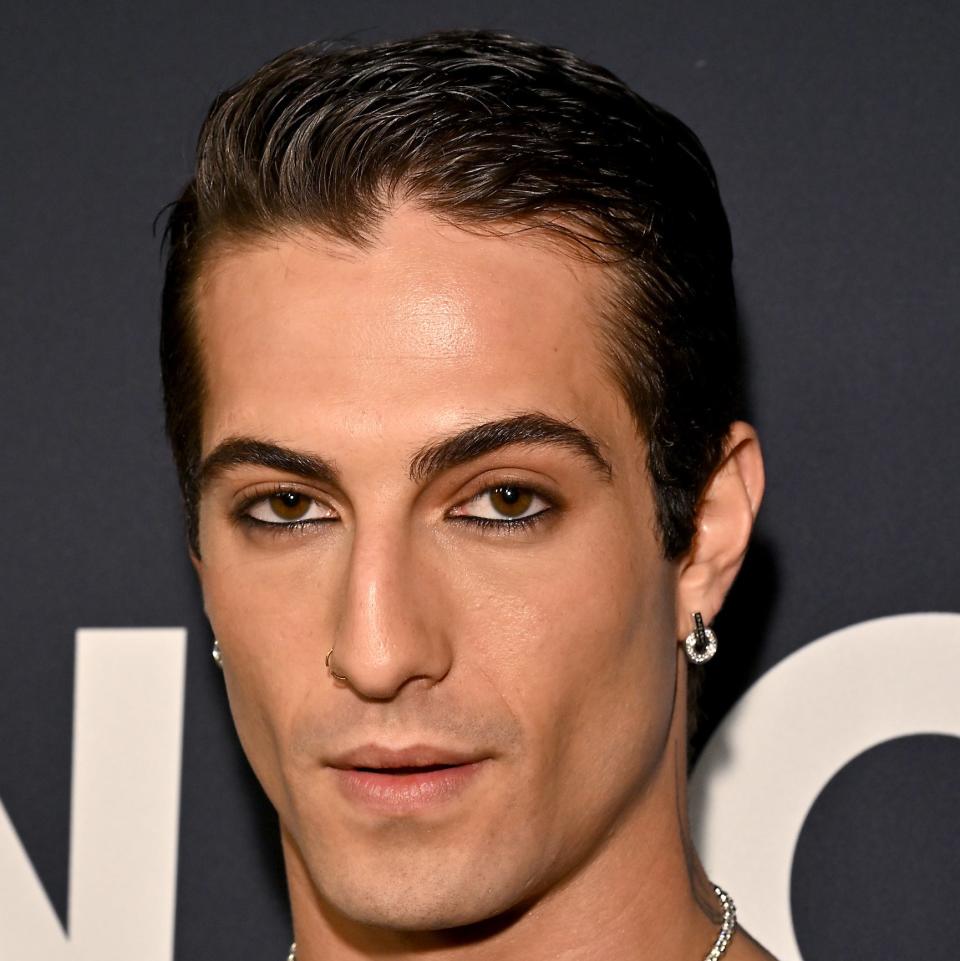 Damiano with slicked-back hair, keyhole cutout top, and tattoos, posing at an event