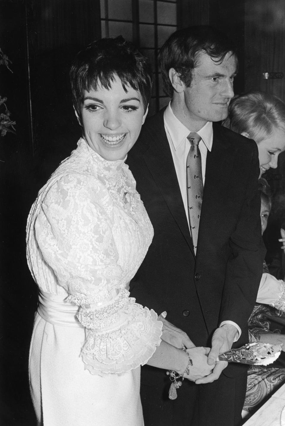 Liza Minnelli, wearing a white lace bridal gown, smiles as she cuts her wedding cake with her husband, Peter Allen.