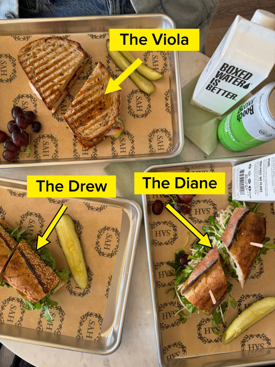 Three sandwiches on trays, each with a pickle and grapes. A container of Boxed Water and a can of drink are on the side