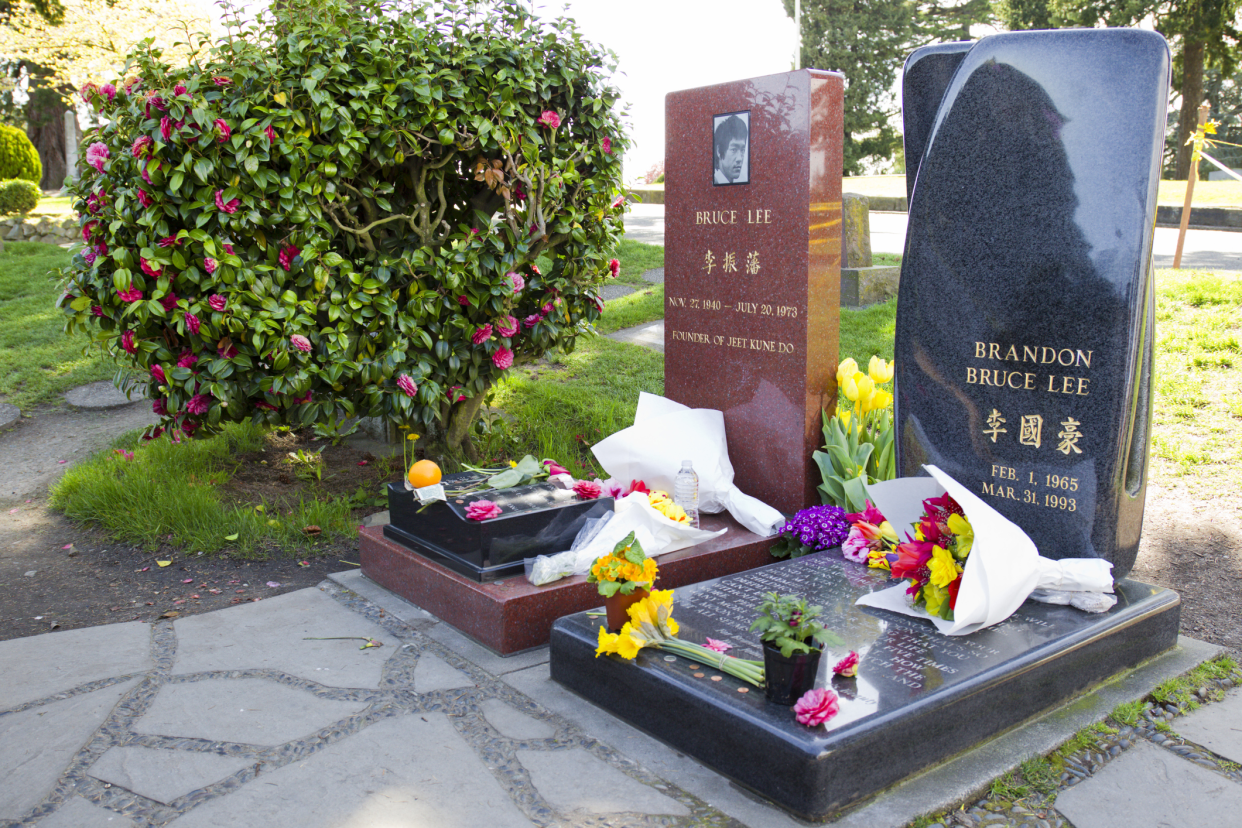 Bruce Lee's grave in the Lakeview Cemetery, Seattle with Brandon Bruce Lee's grave on the right and a pink flower bush on the right with several flowers on the graves, on a sunny day