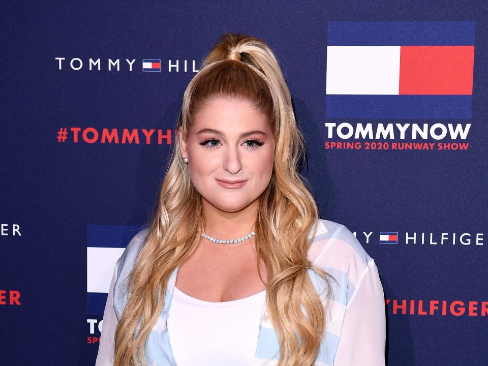 meghan trainor on a tommy hilfiger red carpet, wearing a light blue and white striped dress with a long sleeved white undershirt. her hair is half up in a high ponytail, and softly curled