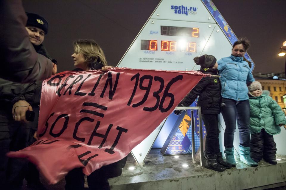 A gay rights activist holds a banner in front of a large clock showing the number of days left until the start of the Olympic games as police officers approach, left, in St. Petersburg, Russia, Wednesday, Feb. 5, 2014. Russian gay rights activists protested the upcoming Olympic Games in Sochi. Two activists unfurled banners reading “Berlin 1936 = Sochi 2014,” referring to the Olympic Games that were held in the capital of Nazi Germany. One-man pickets are legal in Russia and the two activists holding signs were spaced far enough apart that neither was arrested. (AP Photo/Elena Ignatyeva)