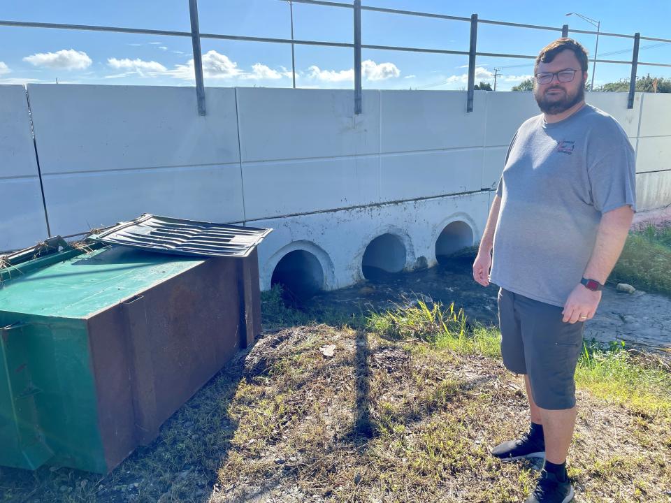 Advanced Roof Technology Office Coordinator Brandon Massimini stands Monday morning alongside a green metal trash bin that floated onto his company's lot. The three drainage culverts in Brightline's concrete retaining wall were overwhelmed by floodwater.