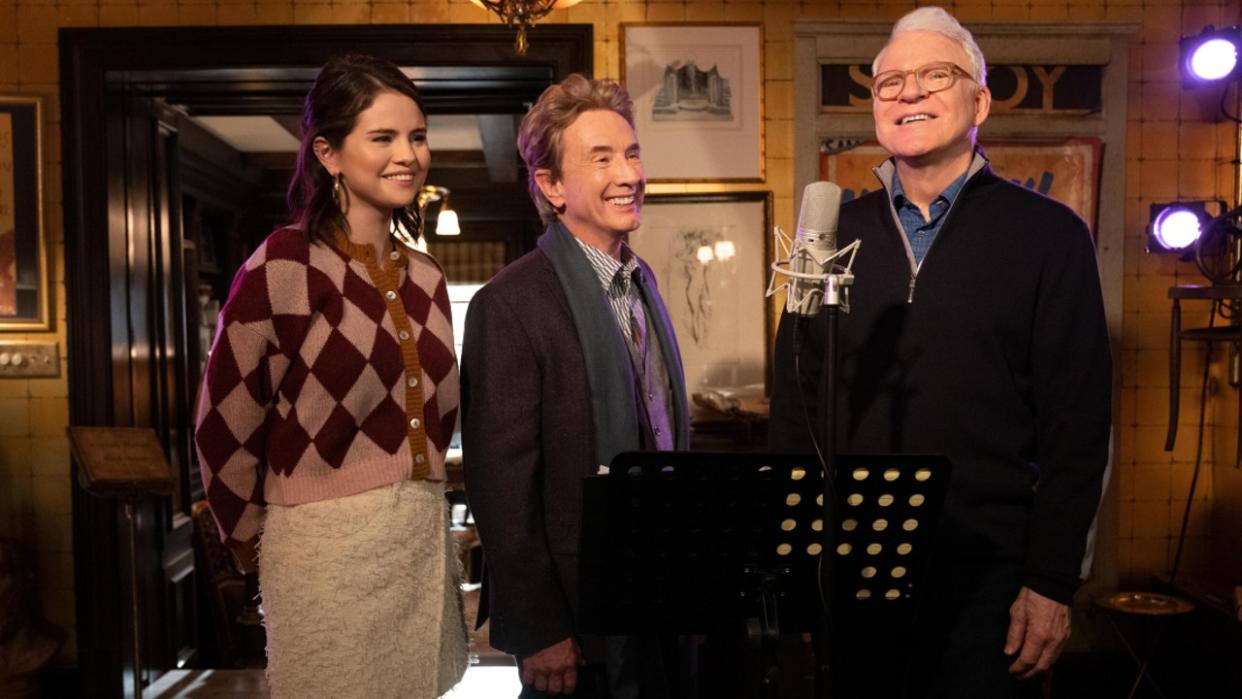  From left to right: Selena Gomez, Martin Short and Steve Martin standing around a microphone recording their podcast. 