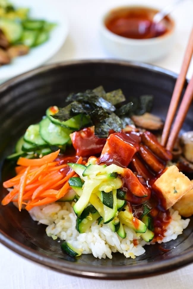 A close-up of a rice bowl topped with sliced vegetables, seaweed, tofu, and a red sauce. A small dish with a spoon of red sauce is in the background