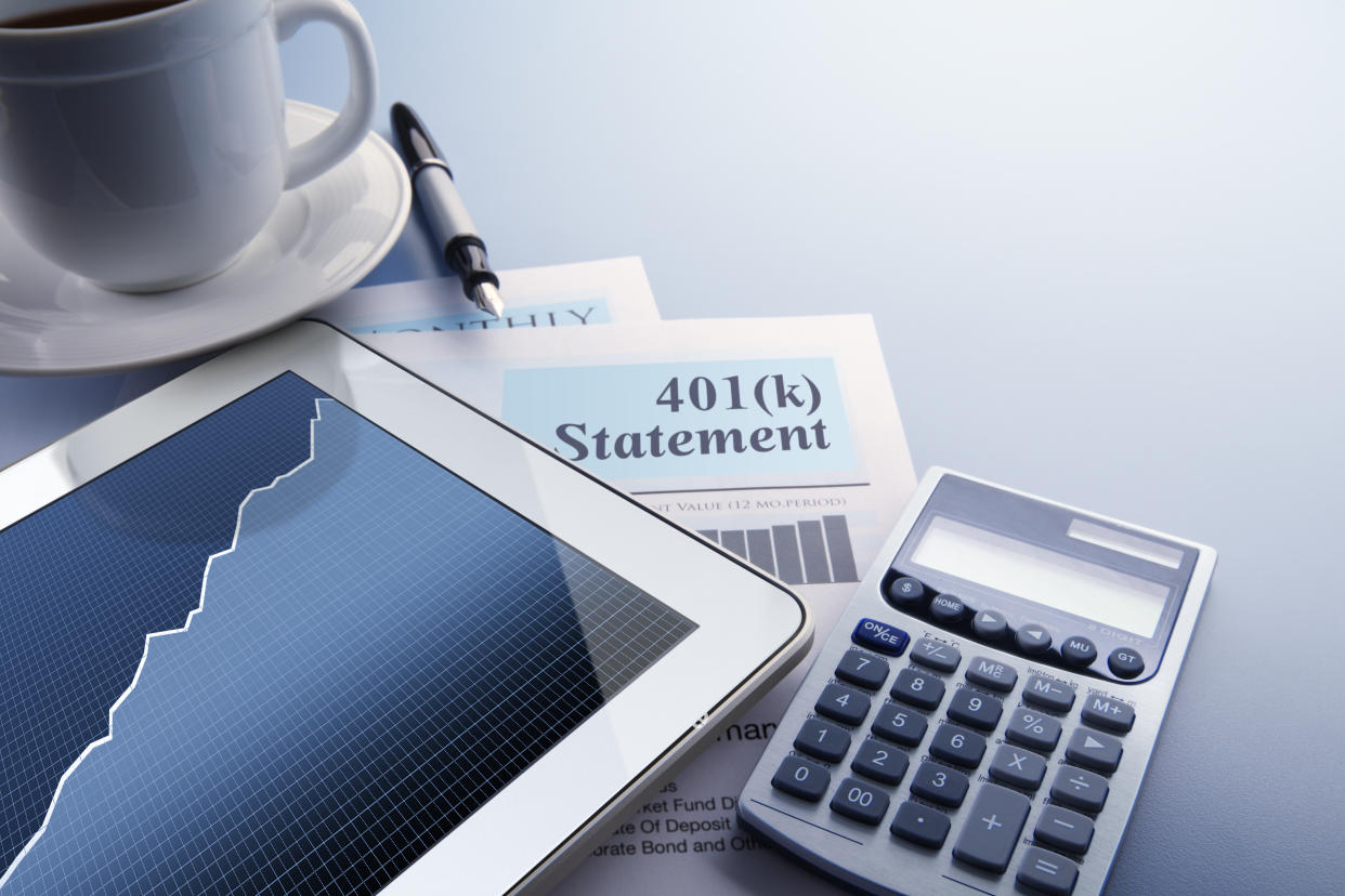 A digital tablet with a stock chart on the screen rests on top of a 401(k) statement on a desktop. A fountain pen, a calculator and a cup of coffee also frame the scene.