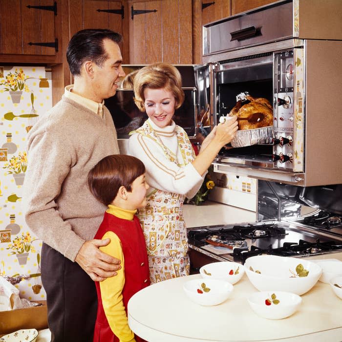 A man, woman, and young boy stand in a kitchen. The woman is taking a cooked turkey out of the oven while the man and boy watch. The scene appears to be from the mid-20th century