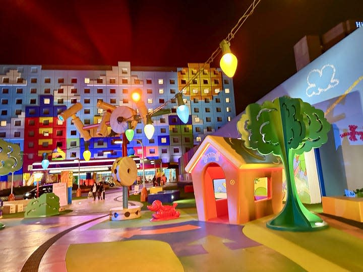A courtyard at the Toy Story Hotel in Tokyo is shown at night. A dog house, toy trees, and other oversized toys are visible.